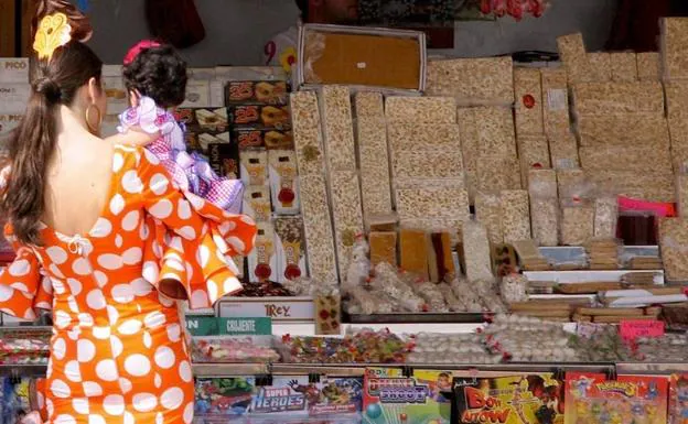 Una mujer con traje de flamenca, con una niña en brazos, frente a un puesto ambulantes de dulces y turrones. /
